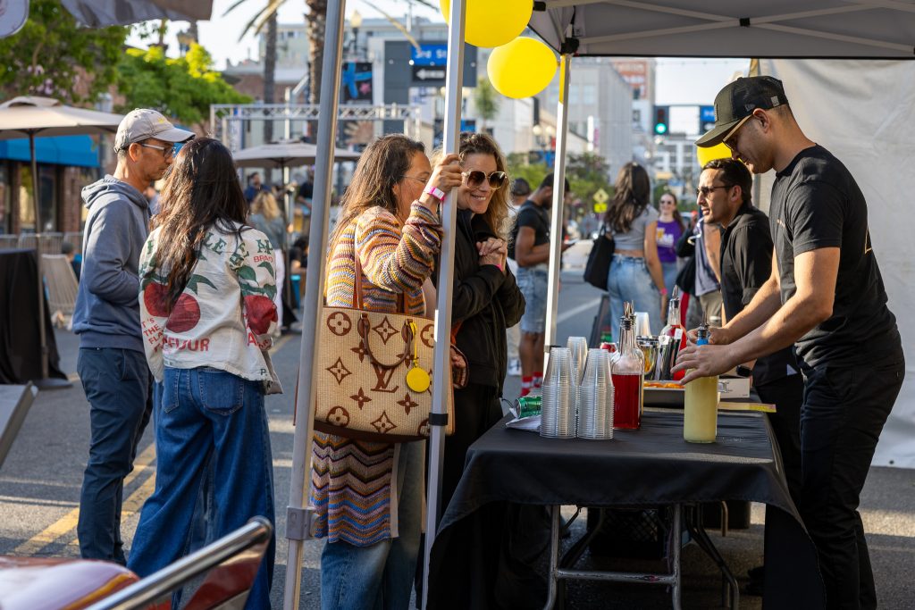 Two women chat at a street festival beside a beverage stand under a canopy with yellow balloons nearby.