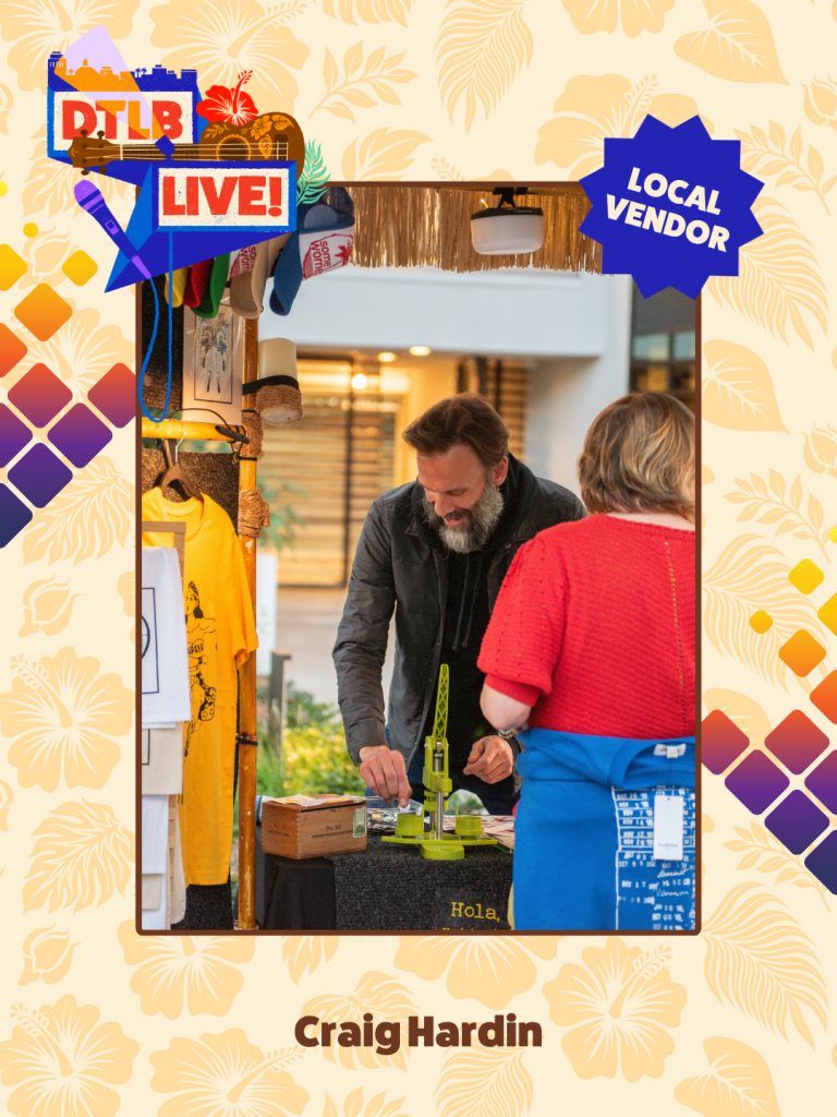 Man with beard at a vendor booth showing a green hand-crank tool to a woman in a red top; 'Local Vendor' badge visible in the background.