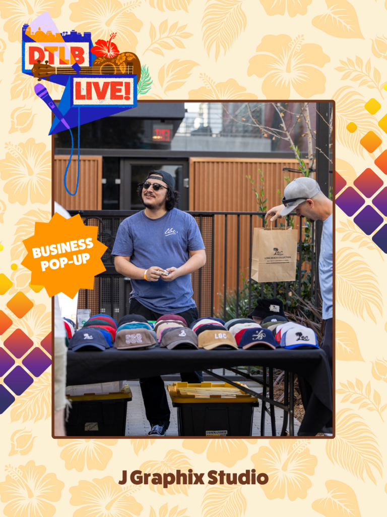 Outdoor street market scene featuring a man in a blue t-shirt behind a table of colorful baseball caps; another person with a shopping bag nearby. Decorative signs read 'Business Pop-Up' and 'DTLB Live!' in the top-left corner. The frame includes a tropical patterned border and 'J Graphix Studio' at the bottom.