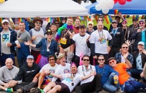 A large, diverse group of people at a Pride event under tents, wearing rainbow accessories and costumes, posing and cheering together.